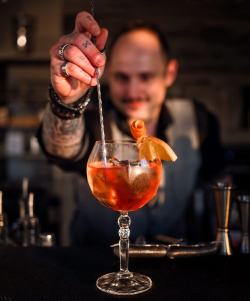 A close-up of a professional barman at Tenuta d’Arbia Boutique Hotel carefully stirring a vibrant Spritz cocktail garnished with a fresh citrus twist. The warm, low-light ambiance of the bar reflects the elegant evening atmosphere of our estate in the Val d’Arbia.